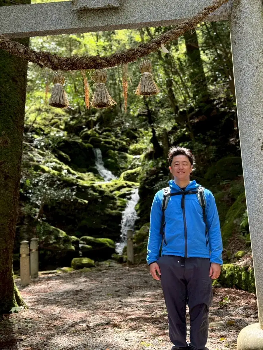 Issei, your guide, standing beneath a stone torii by a mountain waterfall in Tara